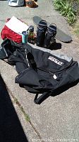 Itech hockey duffel bag laid on concrete with snowboard boots, skateboard deck without wheels, assorted gear including flag football equipment and two sports cups nearby.