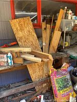 Multiple scrap wood pieces stacked against shelving and an outdoor wall; includes rough and smooth wood, some with nails.