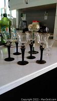 Front view of nine stemware glasses arranged on a white countertop in a kitchen. Five glasses have black twisted stems and gold accents with clear bowls, other glasses include various styles with green stem, black bowl with white design, and clear stems.