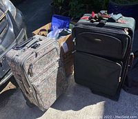 Two travel suitcases and reusable grocery bags placed on pavement beside vehicle. One suitcase floral patterned and smaller, the other dark green and larger.