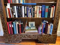 Front view of wooden bookshelf containing two shelves fully loaded with books and binders, lower shelf includes a framed photo and a box with postcards