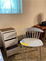 Full view of painted wood chair and chest of drawers beside a bed in a room with natural light from a window. Chair has a curved top rail and spindles and is white painted. Chest has three drawers with round knobs and a dark textured top surface.