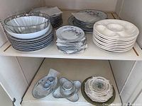 Wide view of dinnerware with stacks of plates, serving trays, and a condiment set on two shelves in a cabinet.