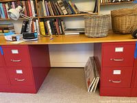 Overall front view of two red metal file cabinets under a wooden desk with books and baskets on top.