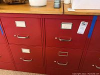 Front view showing two red metal file cabinets side by side with two drawers each. Each drawer has label holders and silver handles. Lot tag visible.