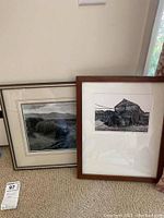 View showing both framed pieces leaning against wall on carpet, one with silver frame and landscape photo, the other with wood frame and print of barn.