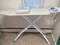 White extra-wide ironing board with iron rest and control box on end, standing on tile floor near curtain and baseboard heater.