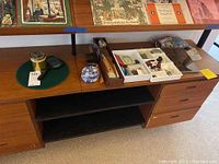 Overall view of the desk shelf showing plastic bins with office supplies, ceramic trinket box, wooden thermometer, keys, and magnifying glass on green cloth.