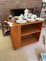 Wooden rolling cart with storage shelves on multiple sides and caster wheels, shown with a set of ceramics on top.