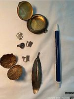Top view of the array of silver plated items: round locket trinket box, clam shell trinket box, two clip-on earrings, two charms, and leaf-shaped pin brooch next to a pen for scale.