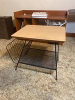 Front-angle side view of the vintage magazine table on carpeted floor showing the wood top, metal frame, mesh shelf, and wire magazine rack.
