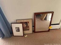 Three framed pieces leaning against wall: rectangular wood mirror, silver-framed print, wood-framed portrait
