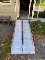 Top view of metal foldable wheelchair ramp laid out on outdoor patio, showing hinge connection and ribbed surface for traction. Shows some dirt and leaf debris on surface.