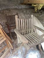 Three solid wood deck chairs in weathered condition stored in an unfinished area beneath a house, showing slatted wooden seats and backs, with curved wooden armrests and legs.