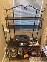 Full front view of wrought iron bakers rack with three black shelves and decorative leaf and acorn design on top and sides. Window blinds background.