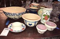 Photo showing a variety of pottery items including a large cream-colored Marshall Pottery popcorn bowl with blue rim and text, several pitchers in various colors and patterns including English Torquay motto ware, Bauer, and Czecko Slovakian Erphila, small cream bowls with pink interiors, and other pottery pieces, arranged on a wooden surface.