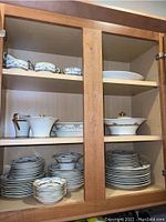Cabinet housing multiple pieces of the Theodore Haviland china set including plates, bowls, serving pieces, and cups seen stacked and arranged on shelves.