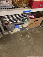 Plastic bin filled with folded Christmas linens including checkered blue and white pieces and red patterned linens, alongside an adjacent cardboard box.