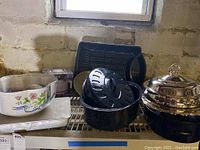Shelf showing multiple cookware and serving items including black enamelware roaster, covered silver plated pieces, and decorative ceramic bowl.