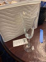 Single crystal champagne flute glass shown on dark wooden table with price tag visible, next to a beige textured fabric storage bin.