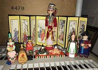 Full view of seven ethnic dolls placed in front of a folding screen with painted figures, showing varied traditional costumes and cultural representation.
