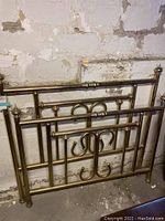 Full view of the brass-like metal headboard and footboard set leaning against a stone wall, showing the entire frame and structure.
