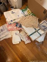 Assortment of Irish linens including folded tablecloths, napkins, and a crocheted doily placed on the floor near a cardboard box.