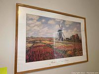 Framed wall art hanging on a white wall, showing a landscape of tulip fields and a windmill under a cloudy sky, with natural wood frame.