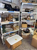 Two white plastic shelving units with various items on them. The items are excluded from the lot. Shelving units show dirt and are located in a basement.