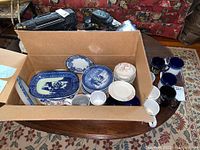 Box holding a mix of plates, bowls, and mugs on a table with patterned carpet background.