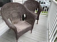 Two brown weathered rattan arm chairs placed on a porch. Visible wear on rattan weave and unraveling at chair legs.