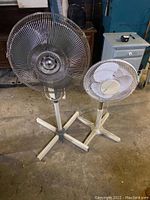 Photo showing two standing fans together in a basement setting, one large with metal grille and one smaller all-white fan.