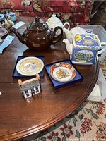 Table with four English tea pots and two decorative plates, showing variety of shapes and colors of teapots including brown, white, scenic, and cottage figurine
