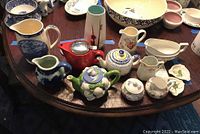 Photo showing multiple ceramic items including teapots, cream and sugar sets, a vase, and small dishes in various colors and patterns on a round wooden table.