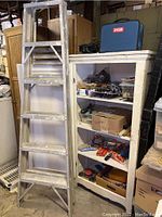Overall lot view showing the metal step ladder leaning against a white shelving unit filled with tools and toolboxes, including a Ryobi drill case on top.