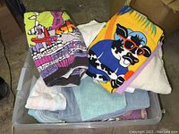 Overview of assorted towels folded in plastic tote, including two printed beach towels and several hand and face towels in solid colors.