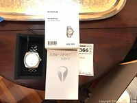 Nixon Monopoly watch in original box with watch outside the box, displaying the silver metal band and white dial. Also includes product paperwork and an instruction leaflet indicating 'New - never worn'.