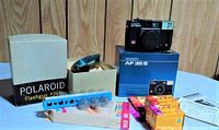 Photo showing the Polaroid Flashgun #268 box, two Kodak 35mm film rolls, a package of Sylvania blue flashbulbs, the Diramac AF35S camera with its box, and two single-use cameras (one new and one used)