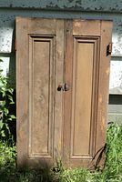 Two very old wooden cupboard doors with visible wear and wooden panel design, placed outdoors against siding over grass.