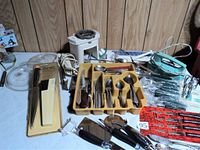 Wide view of all items on table including the yellow cutlery tray, Veg-o-Matic, electric cutter, peelers, knives, and silver pieces.