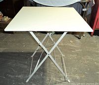 Angle showing rectangular white metal tabletop and foldable cross-braced metal legs against a concrete floor.