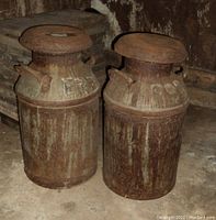 Two old heavily rusted milk cans with lids and side handles sitting on a concrete floor.