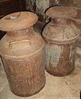 Two heavily rusted metal milk cans placed side-by-side on a concrete floor against a wooden wall.