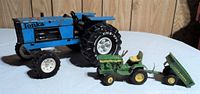 Blue Tonka tractor with rust and white Tonka-branded wheels alongside small green John Deere tractor with dump trailer on white surface