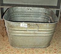 Side view of the old metal wash tub showing its rectangular shape, side handle, and ribbed metal construction with signs of rust and wear.