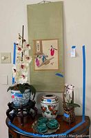 Photo showing full set of Asian décor items arranged including artificial orchids, wall hanging scroll, ceramic jar, and tea set.