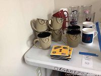 Photo showing 7 beige ceramic coffee mugs with black swirl designs, 2 white ceramic coffee mugs with red and blue prints, and 6 clear plastic Tervis tumblers with logos.