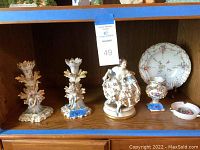 Photo showing two porcelain colonial-style figurines, a decorative plate, a small vase, a small oval dish, and two floral candlesticks with labels showing item numbers