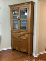 Full front view of the vintage wooden corner cupboard showing upper glass doors with interior shelves, drawer, and lower cupboard doors.