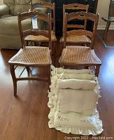 Four wooden dining chairs with carved backs and cane seats shown from an angle. Padded cushions stacked in front.